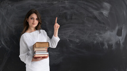 young beautiful girl student stands against the background of a clean chalk board. He holds a stack of textbooks in his hands. The index finger points up.