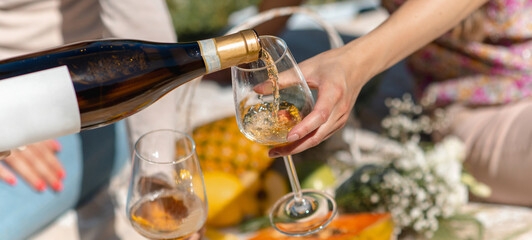 Horizontal banner or header of unrecognizable woman pouring white wine into a glass. Picnic with tropical fruit on background.