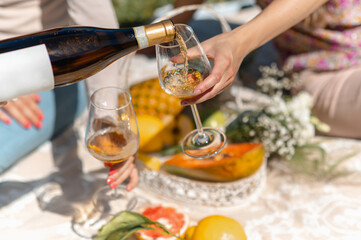 unrecognizable women sitting on a blanket having picnic. Female pouring white wine into a glass. Tropical fruit on background.