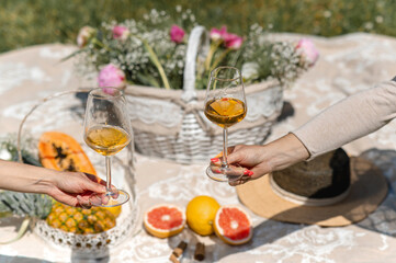 Fototapeta na wymiar Unrecognizable females extending arms to show two glasses with white wine. Blanket picnic on background setting with tropical fruits and flowers.