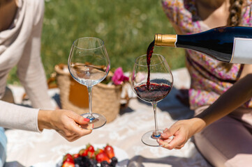Unrecognizable women sitting on a blanket having picnic. Female pouring red wine into a glass....