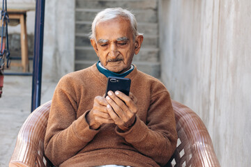 Happy elderly grey-haired Indian man in casual brown clothes using mobile phone, typing sms message. Old people lifestyle concept	