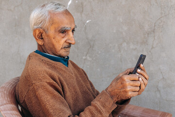 Side view of Senior old Indian man using mobile phone and typing sms, doing online shopping. Modern lifestyle of old people with technology. Copy space in grey background