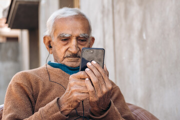 Portrait of old Indian businessman using mobile phone with ear phones. Watching video in mobile. Concept of Technology in elder generation