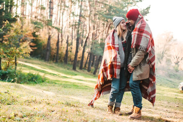 Couple huggings travelers covered with plaid in the forest. Concept of trekking, adventure and seasonal vacation.