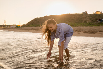 Portrait of beautiful little girl is playing in sea waves in sunset light. Kid and vacation, tourist destination.