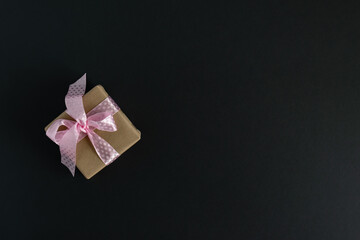 Against a dark background, a gift wrapped in craft paper is tied with a  pink satin ribbon. The concept of the holiday and congratulations. Layout, advertising flyer. Copy-space. Horizontal photo. 