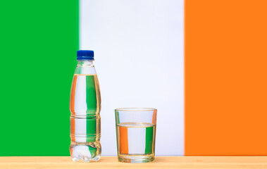 A bottle of clean drinking water and a glass stand on the table against the background of the flag of Ireland. A concept for the supply of clean drinking water in Ireland.