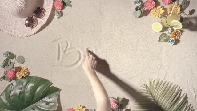 unknown person writing the word beach with his finger on a sandy beach background with a hat, sunglasses, fruit juice, flowers and palm and coconut leaves. concept of summer and holidays.