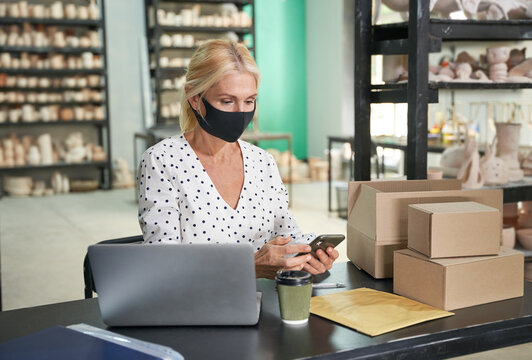 Busy at work. Attractive mature woman, small business owner wearing protective mask using her smartphone while sitting at the desk in her craft pottery shop