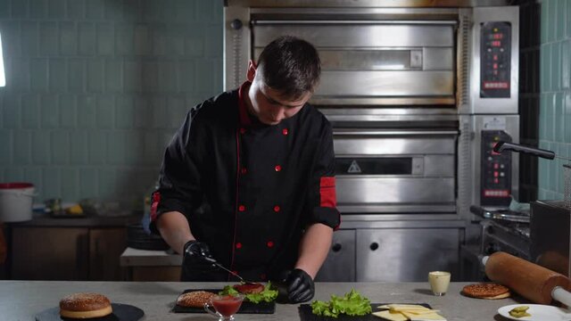 A Caucasian male chef in a restaurant kitchen prepares a hamburger or a cheeseburger dressed in a jacket for cooks. Place the red sauce on the cutlet.