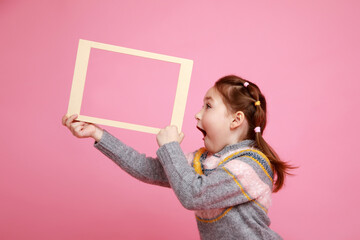 Portrait of a little screaming girl holding blank frame for mock-up on a pink background