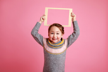 Portrait of a little smiling girl holding blank frame for mock-up on a pink background