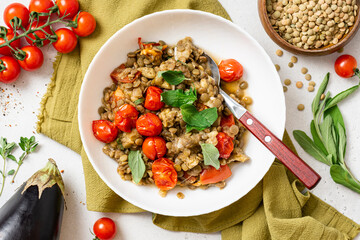 Stewed green lentils with tomatoes, eggplants, spices, and herbs in a white plate on a gray concrete background top view. Vegan food. Lentils with vegetables.