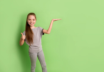 Little teenager smiling girl is showing empty copy space on her open hand with thumb up on other hand on green background in studio.. Happy kid proposing a product. Copy space