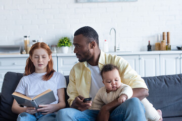 african american man holding tv remote controller near daughter sitting with book with closed eyes