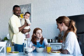 african american man holding infant near wife with daughter having breakfast in kitchen