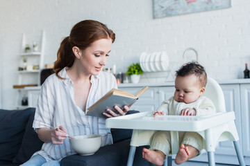 mother reading book to african american baby girl during breakfast