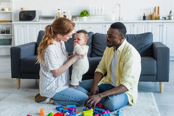 mother holding infant daughter while sitting on floor near african american husband and colorful building blocks