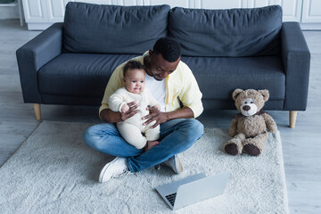 african american man holding baby while sitting on floor near laptop and teddy bear