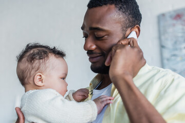 smiling african american man talking on smartphone while holding infant daughter