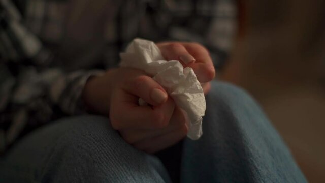 Close-up hands of unrecognizable crying woman holding paper tissues and nervously rummaging through them. Depressed upset female crying alone sitting on bed in dark bedroom. Shooting in slow motion.