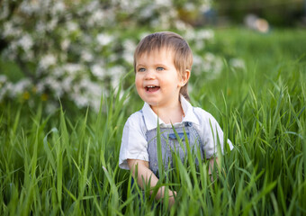 Cute boy in fashionable clothes with blue eyes plays and jumps in the tall grass in a large green blooming garden