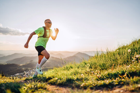 Middle-aged mountain trail runner man dressed bright t-shirt with a backpack in sports sunglasses endurance running uphill by picturesque hills at sunset time. Sporty active people concept image.