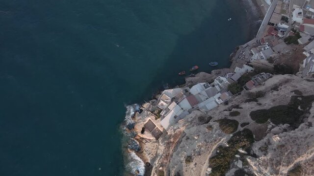 Drone shot of low-rise houses built on a rock by the sea with moored caiques. Aerial view of fishing boats at calm sea in Crete Island at coastal with calm waters.