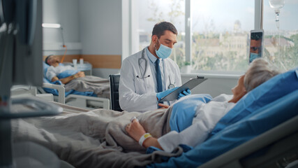Obraz premium Hospital Ward: Friendly Doctor Wearing Face Mask Talks to Beautiful Senior Female Patient Resting in Bed, Explains Test Results, Gives Recovery Advice. Physician and Old Lady Recovering after Covid-19