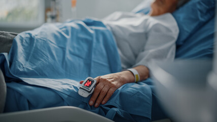 Obraz na płótnie Canvas Hospital Ward: Senior Woman Resting in a bed with Finger Heart Rate Monitor / Pulse oximeter showing Pulse. Her Fragile Hands Resting on a Blanket. Focus on the Hand.