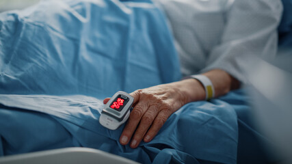 Hospital Ward: Senior Woman Resting in a bed with Finger Heart Rate Monitor / Pulse oximeter showing Pulse. Her Fragile Hands Resting on a Blanket. Focus on the Hand.