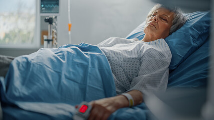 Hospital Ward: Senior Woman Resting in a bed with Finger Heart Rate Monitor / Pulse oximeter...