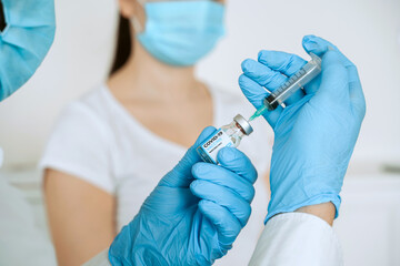 Hand of medical staff in blue glove holding covid-19 vaccine bottle, ready to do injection to woman for coronavirus immunization.