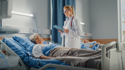 Hospital Ward: Friendly Doctor Asks Health Care Questions of Smiling Elderly Lady Resting in Bed. Physician Uses Tablet Computer, Does Checkup, Talk to Old Woman Fully Recovering after Sickness