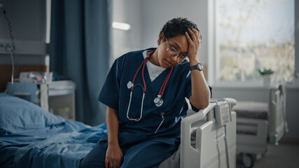 Hospital Ward: Portrait of Sad, Tired Black Nurse Sitting on a Bed, Holding Her Head in Sorrow for...