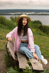 Relaxed beautiful girl in straw boater hat sits on a wooden bench on a background of the picturesque landscape.
