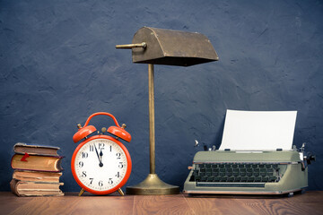 Vintage typewriter with paper blank, orange alarm clock, old desk lamp, aged books on wooden table front concrete black wall background. Retro style filtered photo
