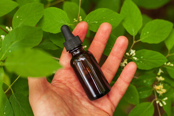 Hand holding a dropper amber bottle containing organic essential oil with leaves in the background