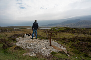 Fototapeta premium Caucasian man with small black and white dog looking out over Loch Druidibeag nature reserve in South Uist. Taken from behind. Rock, fence, loch, grass, mountains in view. Dramatic clouds. 