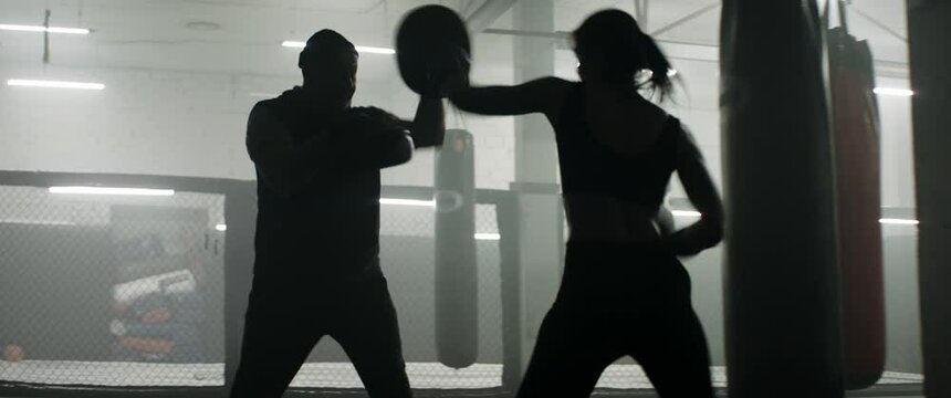 HANDHELD WIDE to CU Caucasian female boxer practicing with her trainer in a boxing gym, preparing for a fight. Shot with 2x anamorphic lens