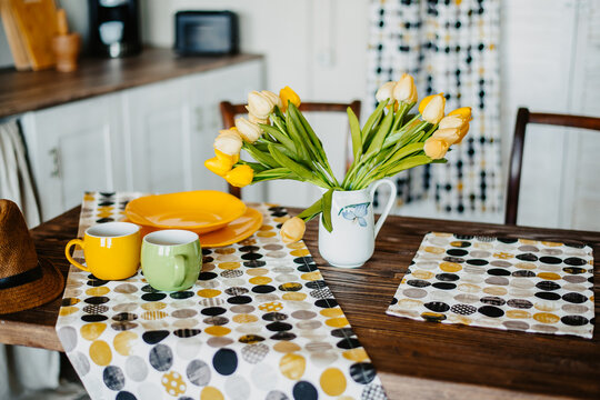 A real photo of a kitchen table covered with a bright polka dot tablecloth and colored bright dishes. On a wooden table there are yellow flowers and tulips. - Powered by Adobe