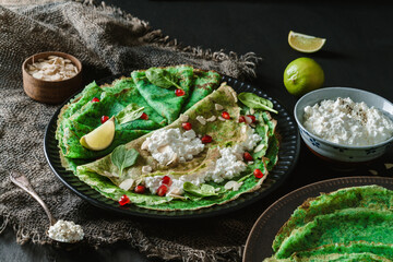 Green vegan crepes with spinach or pancakes with cottage cheese, pomegranate, nuts and spinach leaves on black plate over dark wooden background. Healthy vegan food, homemade breakfast, close up