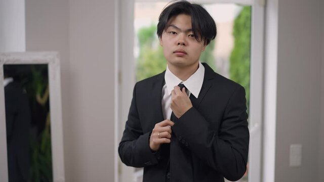 Portrait of focused handsome Asian young groom tying tie looking at camera. Medium shot of concentrated man in elegant wedding suit with boutonniere getting dressed in the morning