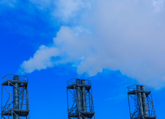 Industrial chimneys with white, thick pollution-causing smoke against a blue sky.