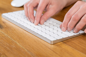 An image of a woman's hand is typing on a white keyboard on a wooden table. Selective focus