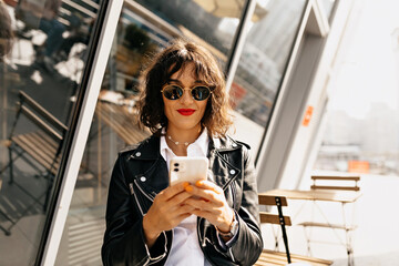 Modern stylish woman with short hairstyle wearing sunglasses using smartphone outdoor in sunlight