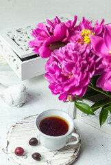 Fototapeta na wymiar Shabby chic tea. A cup of tea on a white wooden tray with a cherry nearby. Peonies are in a white box. Blurred background