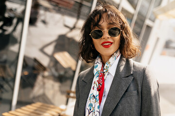 Incredible stylish girl with red lips wearing sunglasses, red jacket and colorful scarf smiling at camera outdoor in sunlight 