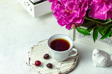 Shabby chic tea. A cup of tea on a white wooden tray with a cherry nearby. Peonies are in a white box. Blurred background